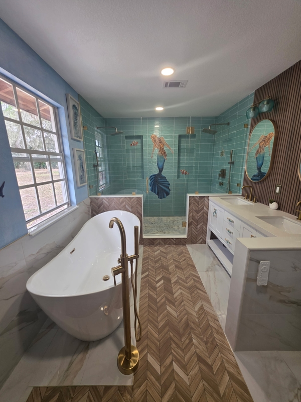 Luxurious bathroom with a white freestanding tub and brass floor faucet, herringbone wood-look floors, teal tiled shower with mermaid mosaic, and a white double vanity.