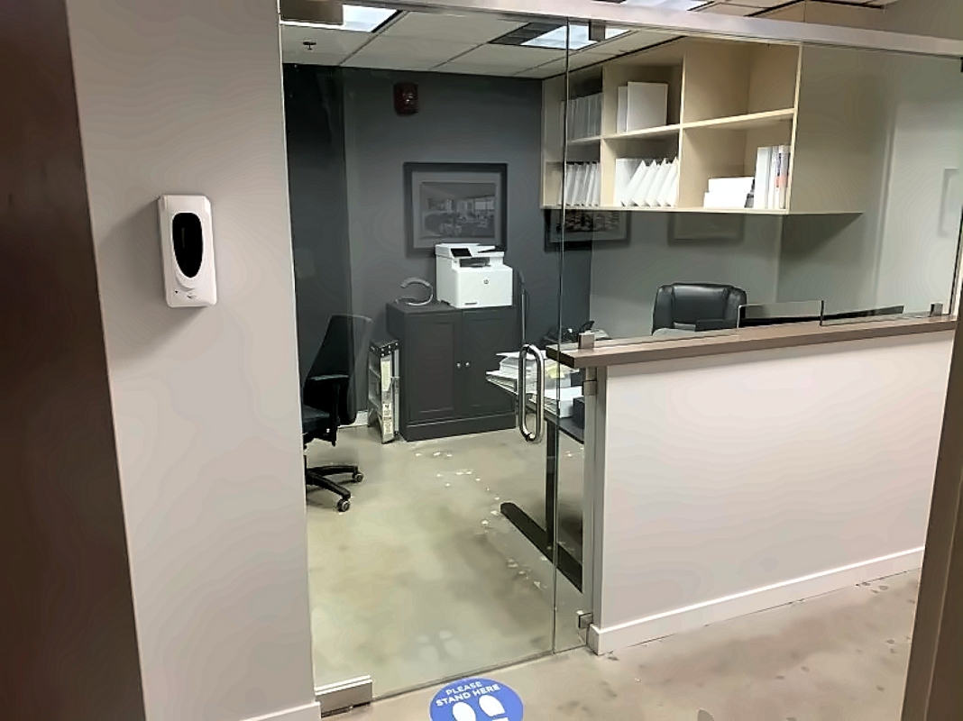 Reception area behind a glass partition with a front desk, a black chair, and shelves; a printer sits on a cabinet in the back.