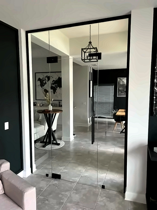Glass-framed double doors open into a modern living area featuring a geometric chandelier, a round pedestal table, wall art, and light gray tiled floors.
