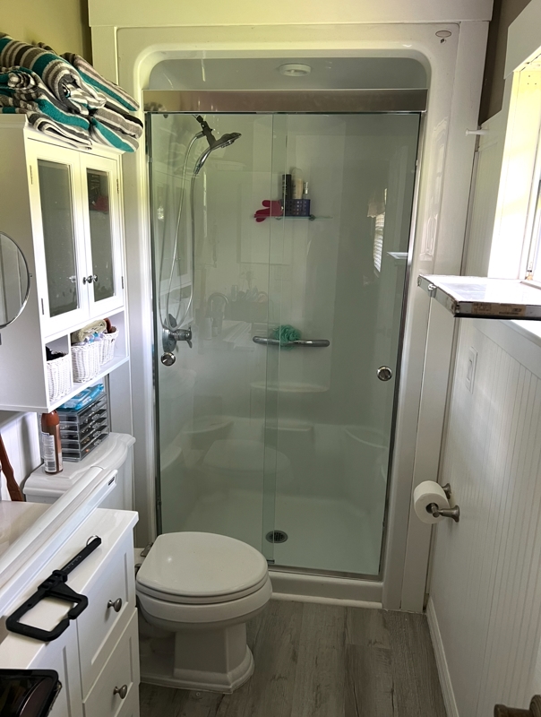 Small bathroom with a glass-enclosed shower, toilet, and white storage cabinet; towels on top shelf.