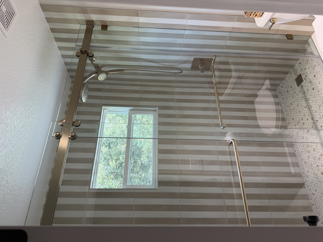 Glass-enclosed shower with striped gray tiles, two grab bars, and a rainfall/he handheld showerhead; small window above a toilet visible outside the enclosure.