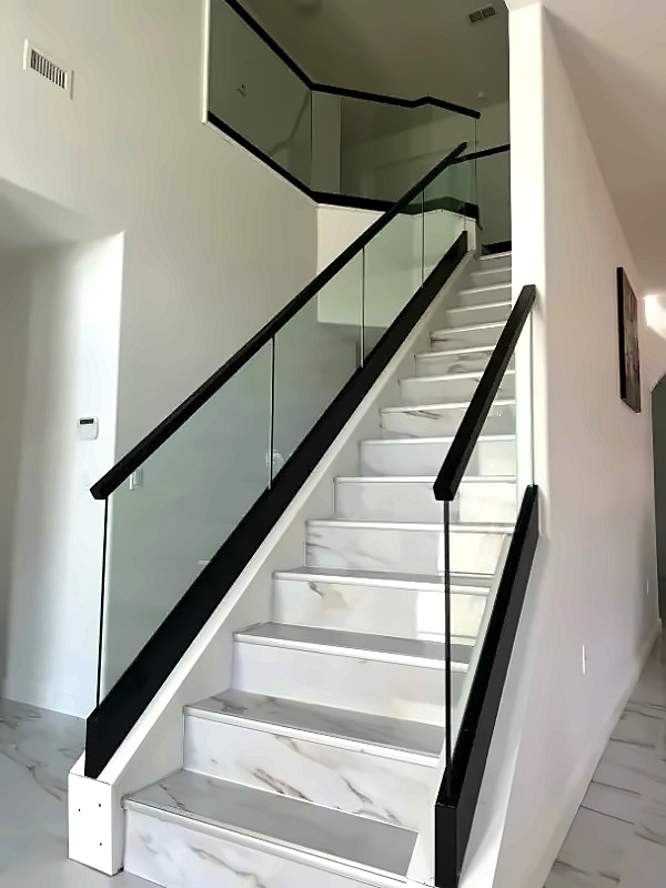 Modern interior staircase with white marble steps, glass balustrade, and black handrails, ascending to a mezzanine in a bright home.