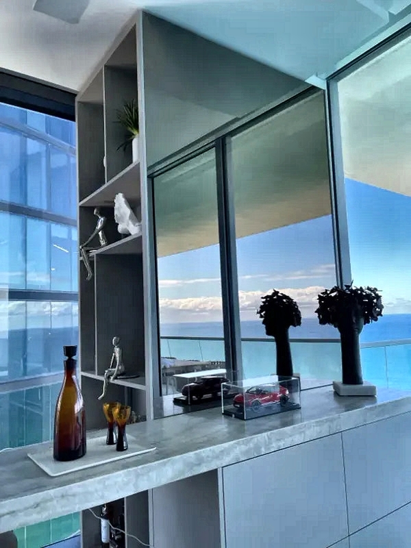 Contemporary interior with marble counter and open shelving, a bottle with two glasses, model cars in a glass case, decorative plants, and large windows showing an ocean view.