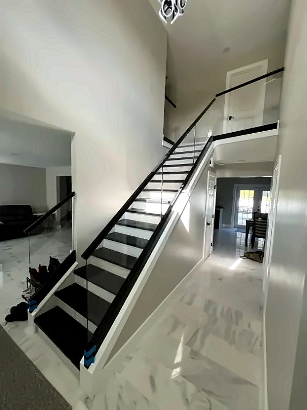 Open two-story foyer with a diagonal staircase featuring a glass railing, black handrails, white walls, and a marble floor bathed in light.