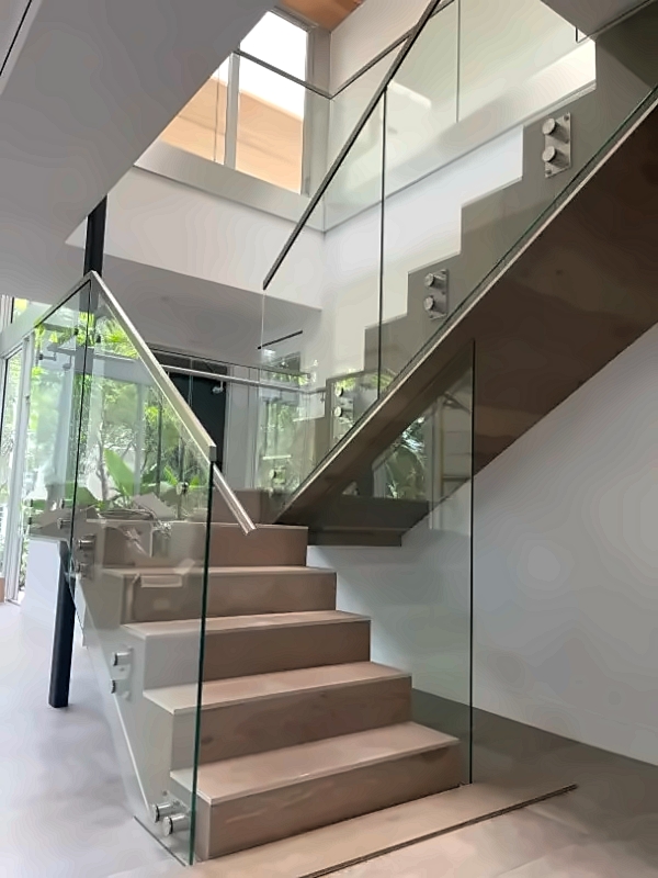 Modern open interior staircase with wooden steps and glass railing, metal supports, sunlight streaming through large windows.