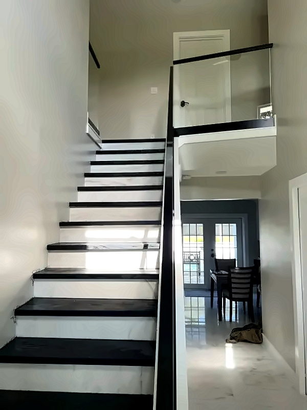 Two-tone staircase with black treads and white risers ascends to a upper landing, right-side glass railing visible, dining area seen through an open doorway below.