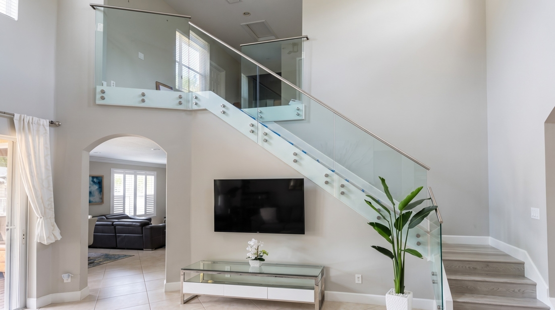 Bright living area with a glass-railed staircase, wall-mounted TV, and a glass console table, plus a tall potted plant near the stairs.